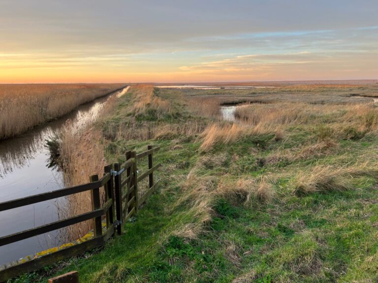 Sunset landscape of Cley-next-the-Sea beautiful marshland nature reserve in Norfolk East Anglia uk on the coast with river water, reeds, fencing and colourful sky on horizon by beach in Winter